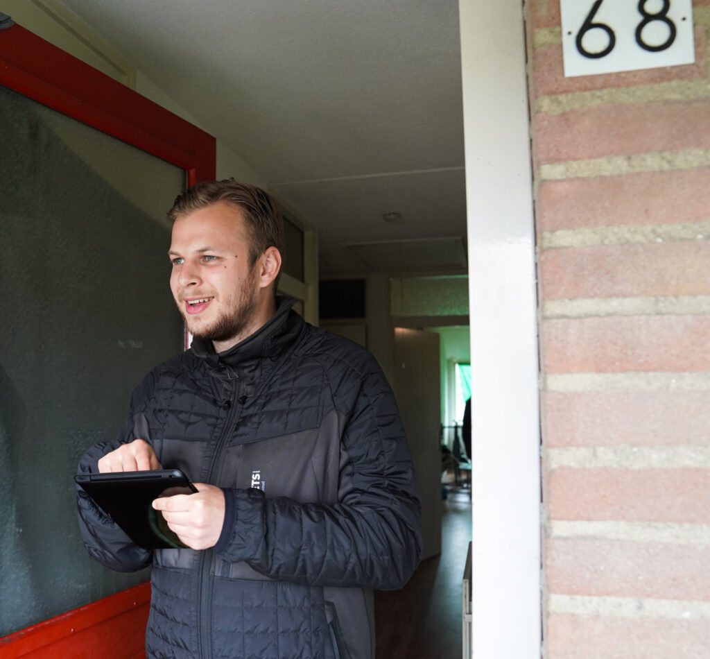 Een jonge man met een tablet in zijn hand bij een bewoner van een woningcorporatie thuis.