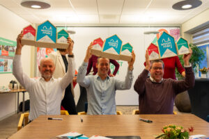 Berrie Endevoets van Orly & Endevoets, Mark Wonders van Woonmeij en Gerard Dinnessen van Caspar de Haan houden houten huisjes omhoog met daarop hen handtekening. Dit symboliseert de ondertekening van het handboek verduurzaming.